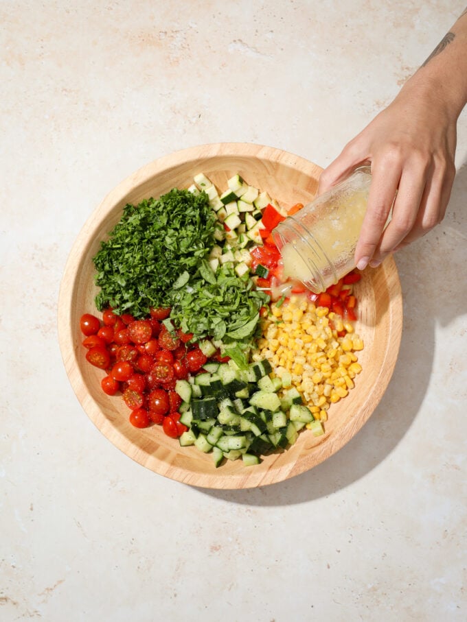 hand pouring dressing over corn, zucchini and tomatoes in bowl